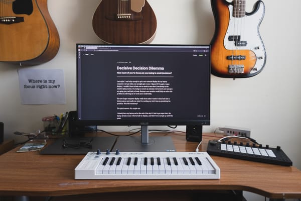 A computer display rests on a desk surrounded by musical instruments, with the words, "Where is my focus?" printed on a piece of paper hanging from the wall.