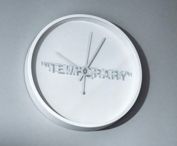 A clock with hands hung on a wall and is photographed with harsh lighting. There are no numbers on the clock. Instead, it has the word temporary written across it.